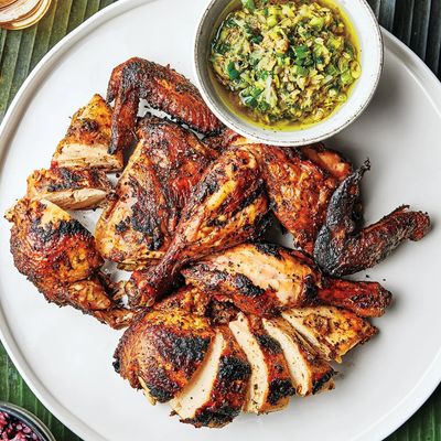 A whole chicken cut into pieces, with char marks from the grill, on a white plate beside a bowl of green sauce